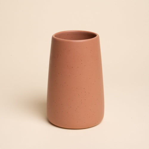 A large minimalist terracotta tumbler vase with subtle iron speckles stands centered on a soft beige background
