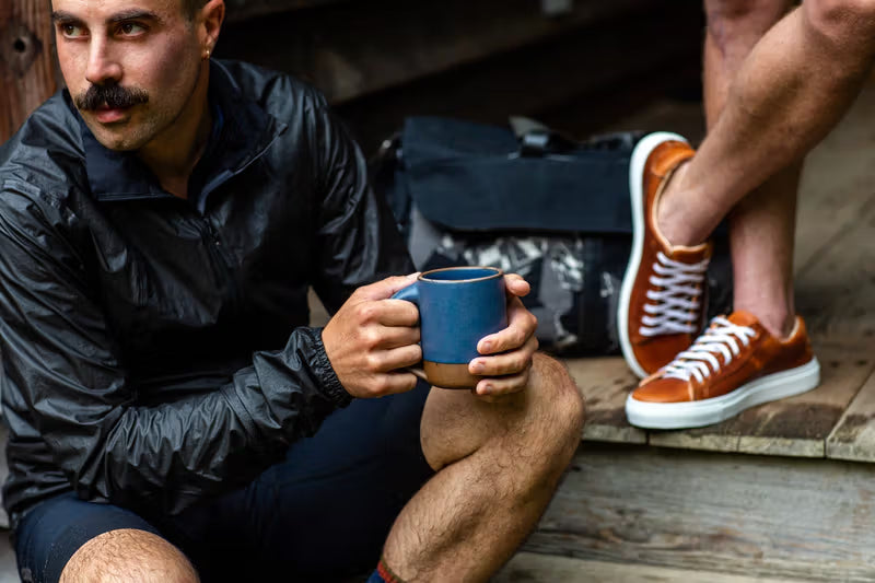 A person seated on a wooden porch holding a blue ceramic mug, wearing a dark jacket. Another person is standing nearby, wearing brown leather sneakers with white soles. A bag is placed in the background.