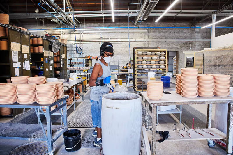 Person in a workshop wearing protective gear is engaged in the pottery-making process, surrounded by stacks of unglazed ceramic plates and pottery equipment on metal tables.