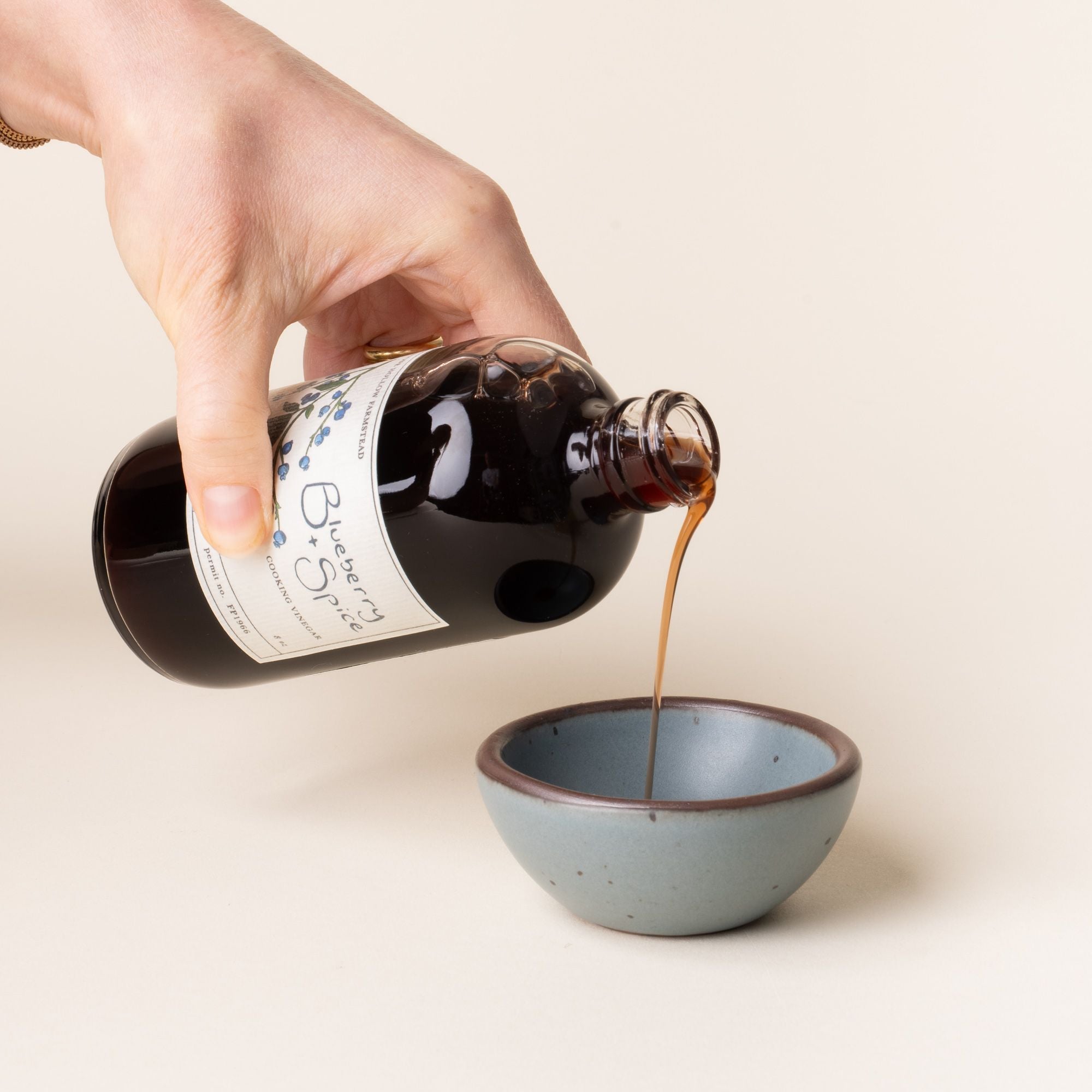 Hand pouring a bottle of Blueberry + Spice cooking vinegar, labeled with hand-drawn blueberry illustrations, into a small ceramic blue-grey bowl.