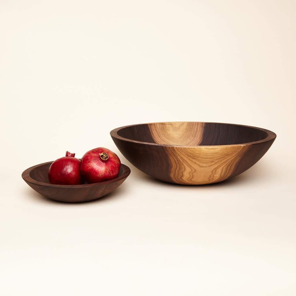 A large-sized walnut wood bowl with some maplewood color in the wood next to a shallow walnut nesting bowl filled with some pomegranates