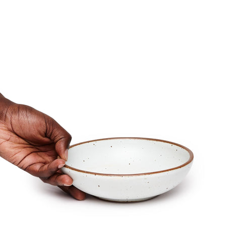 A hand holding a dinner-sized shallow ceramic bowl in a cool white color featuring iron speckles and an unglazed rim
