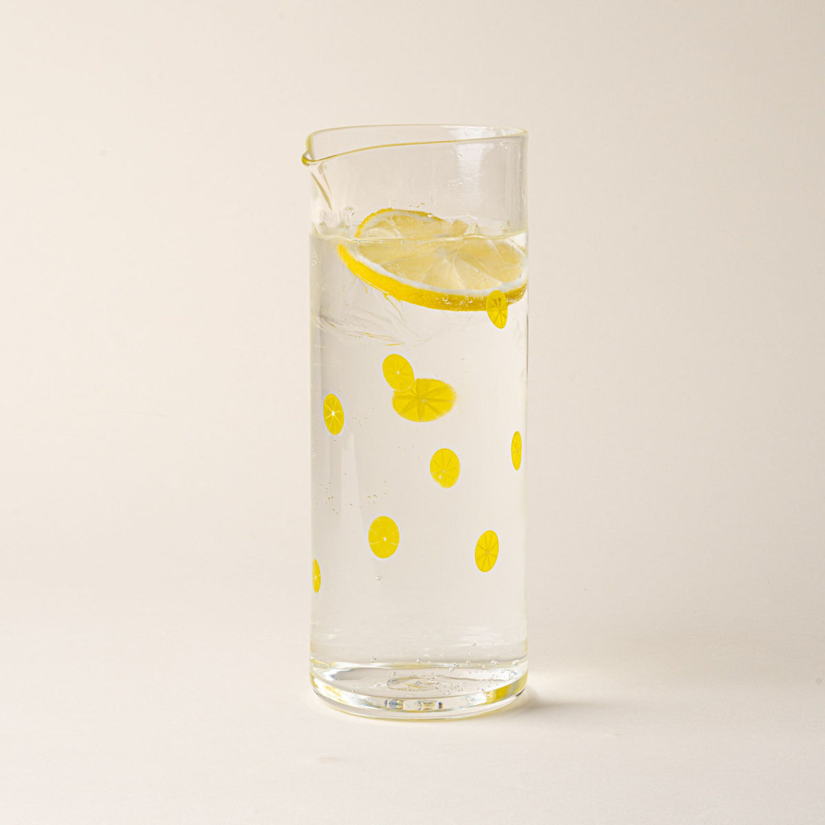 Clear glass pitcher with yellow lemon slice motif and pour spout, filled with water and a slice of lemon.