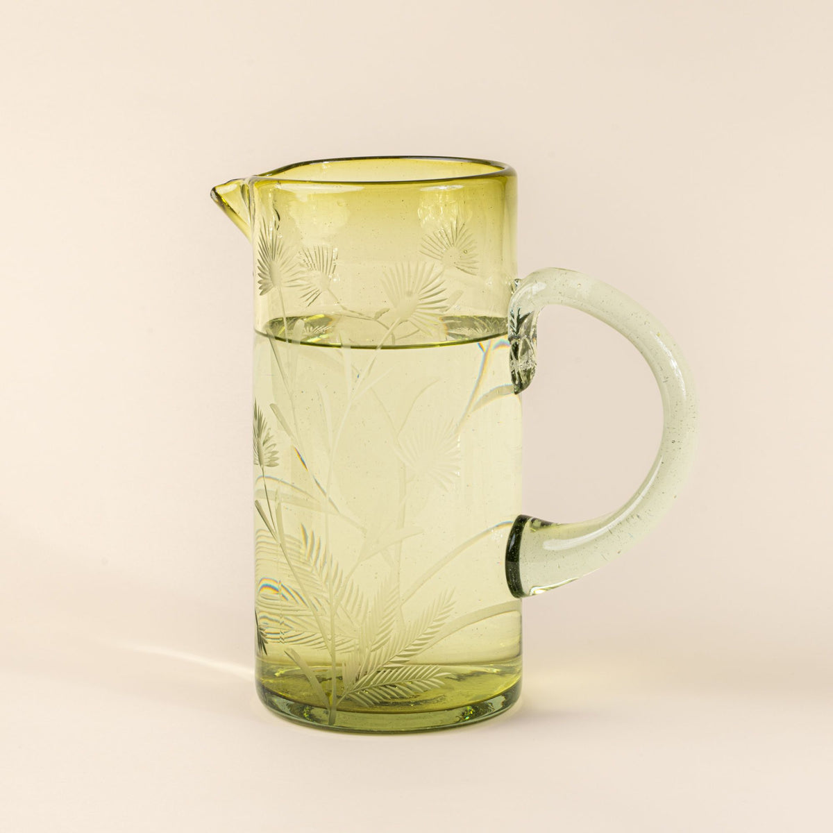 A large glass olive pitcher with handle made of recycled glass with etched thistle designs on it, and filled with water.