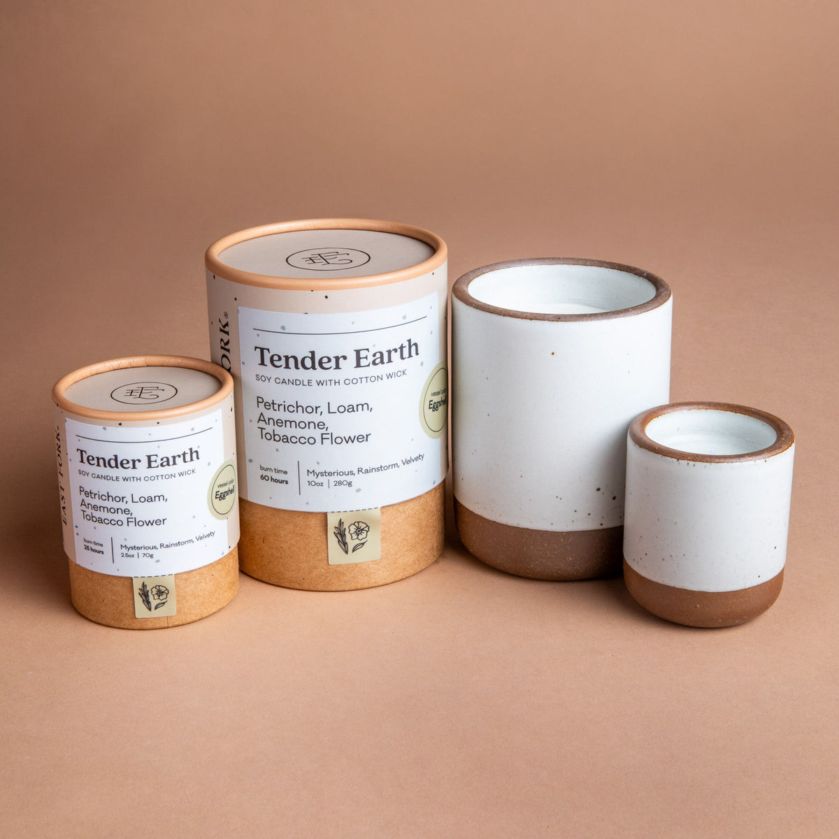 Small and large ceramic vessel next to each other in cool white color with candles inside each. Cardboard tube packaging is on the left with branding stickers that say "Tender Earth".