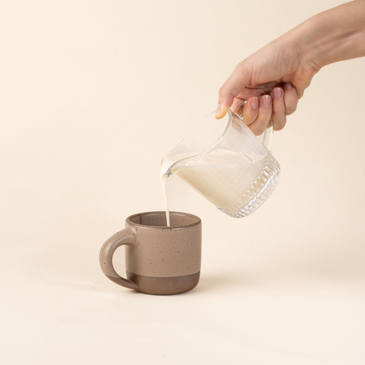 Hand holding a clear glass pitcher with handle and spout on pouring milk into a small ceramic pale warm brown mug.