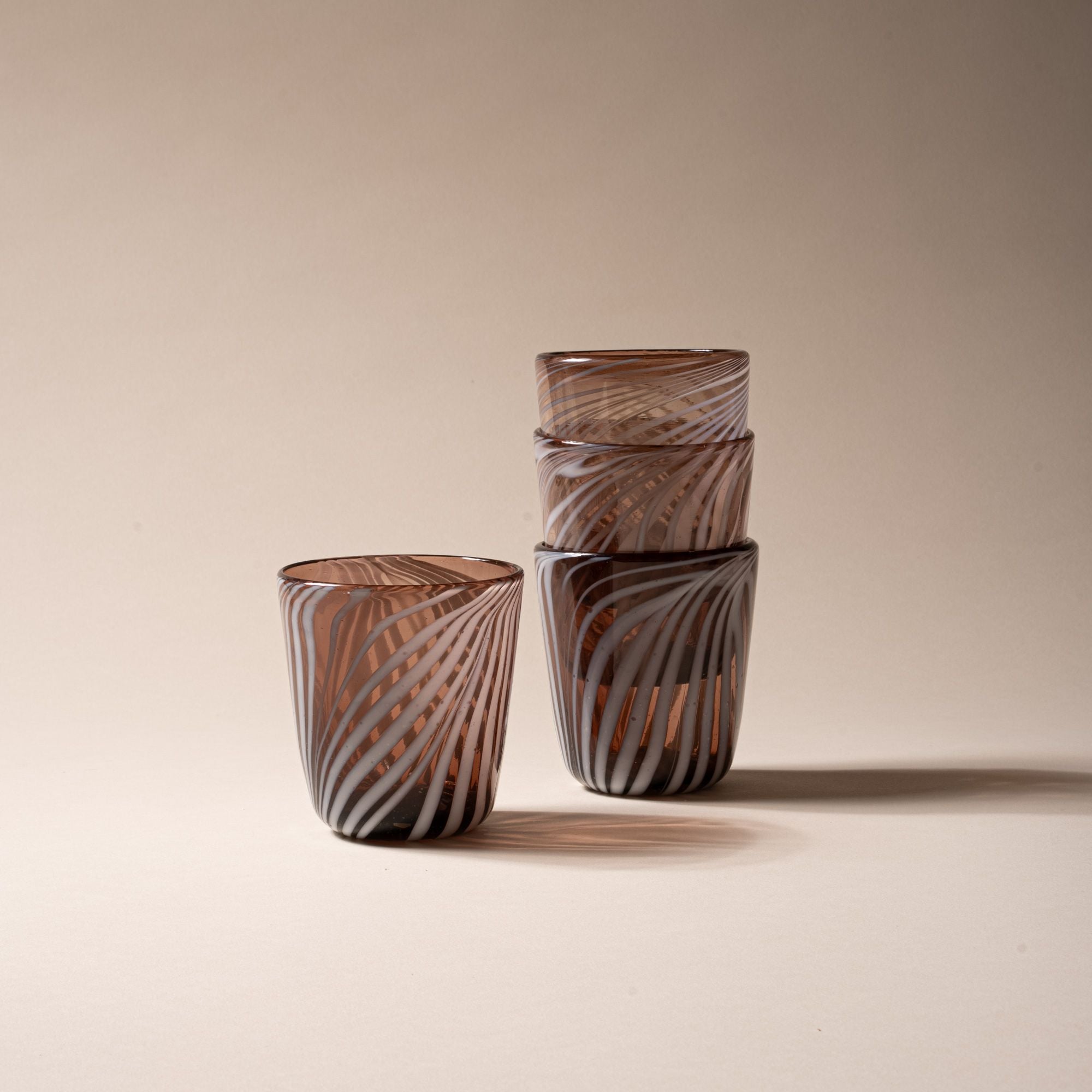 Set of four amber-toned short tumblers with white swirling stripe design, three stacked and one standing beside on neutral backdrop.