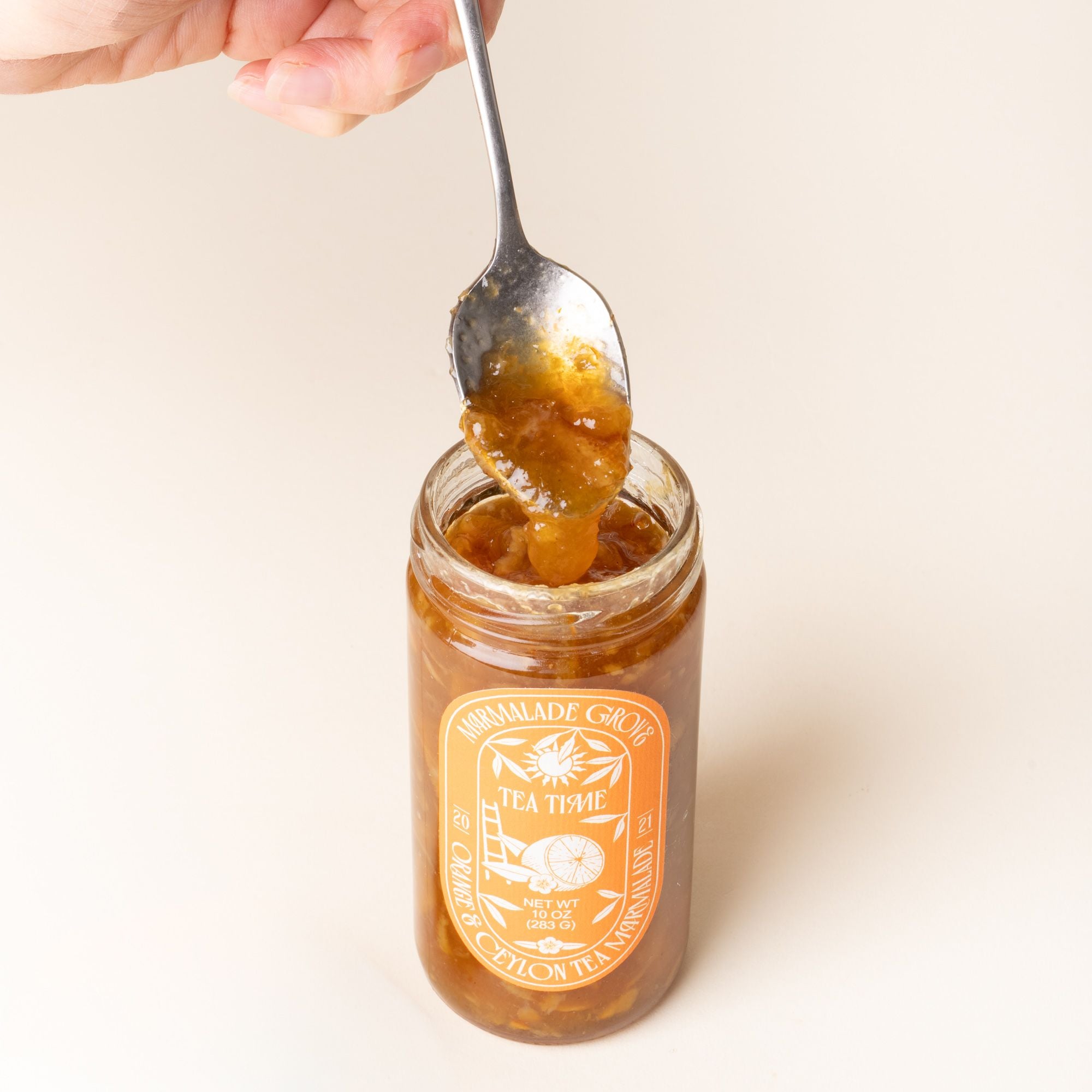 A hand holds a spoon scooping marmalade above an open jar of Marmalade Grove Orange & Ceylon Tea Marmalade, with a bright orange label featuring citrus and floral illustrations.