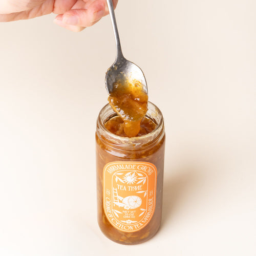 A hand holds a spoon scooping marmalade above an open jar of Marmalade Grove Orange & Ceylon Tea Marmalade, with a bright orange label featuring citrus and floral illustrations.