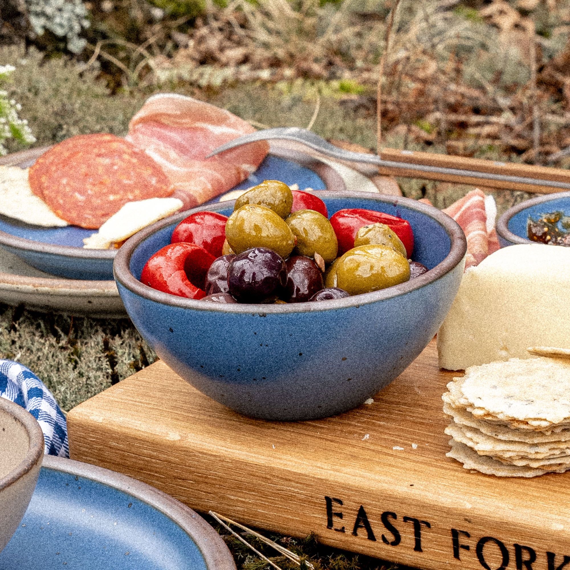 A small dessert sized rounded ceramic bowl in a cool, medium blue color featuring iron speckles and an unglazed rim, filled with olives at an outdoor picnic