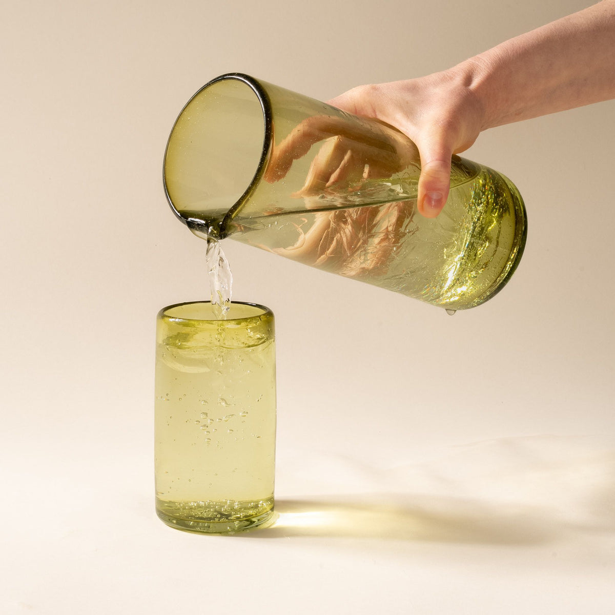 A hand holds an olive green transparent glass pitcher with small spout at the top, and pours a drink into a tall tumbler.