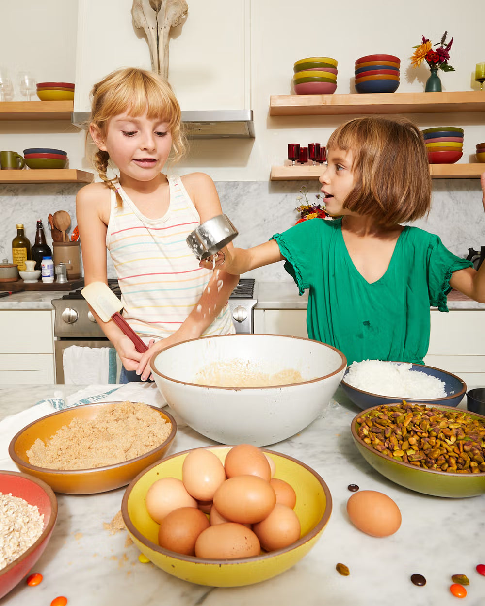 Two children are baking together in a kitchen. The countertop is filled with bowls containing eggs, brown sugar, pistachios, oats, and flour, while kitchen shelves display stacked colorful bowls and glasses.