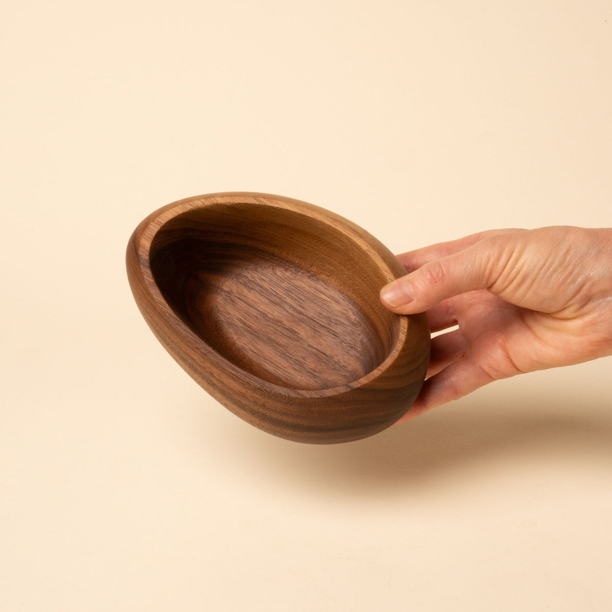 Hand holds out a carved wooden bowl with a smooth, rounded shape and rich brown grain, set against a neutral background.