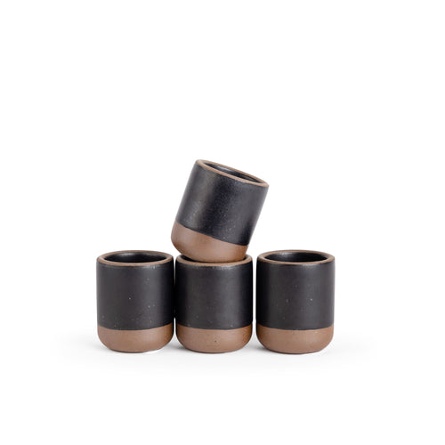 A set of 4 small, short ceramic mug cups in a graphite black color featuring iron speckles and unglazed rim and bottom base, stacked in a row of 3, with the 4th on top.
