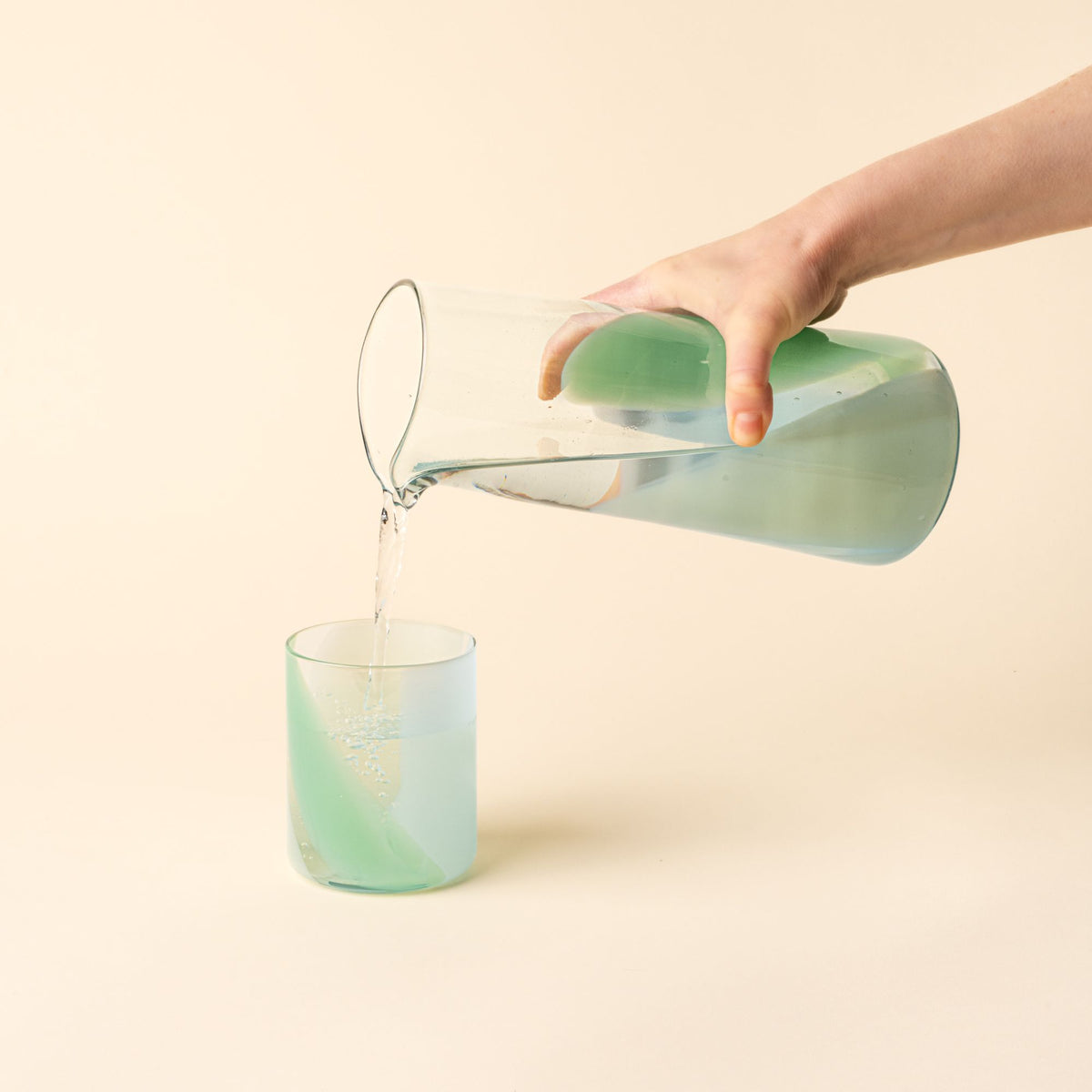 Hand holds a simple glass pitcher featuring a soft pastel green swirl design and pours water into a matching tumbler