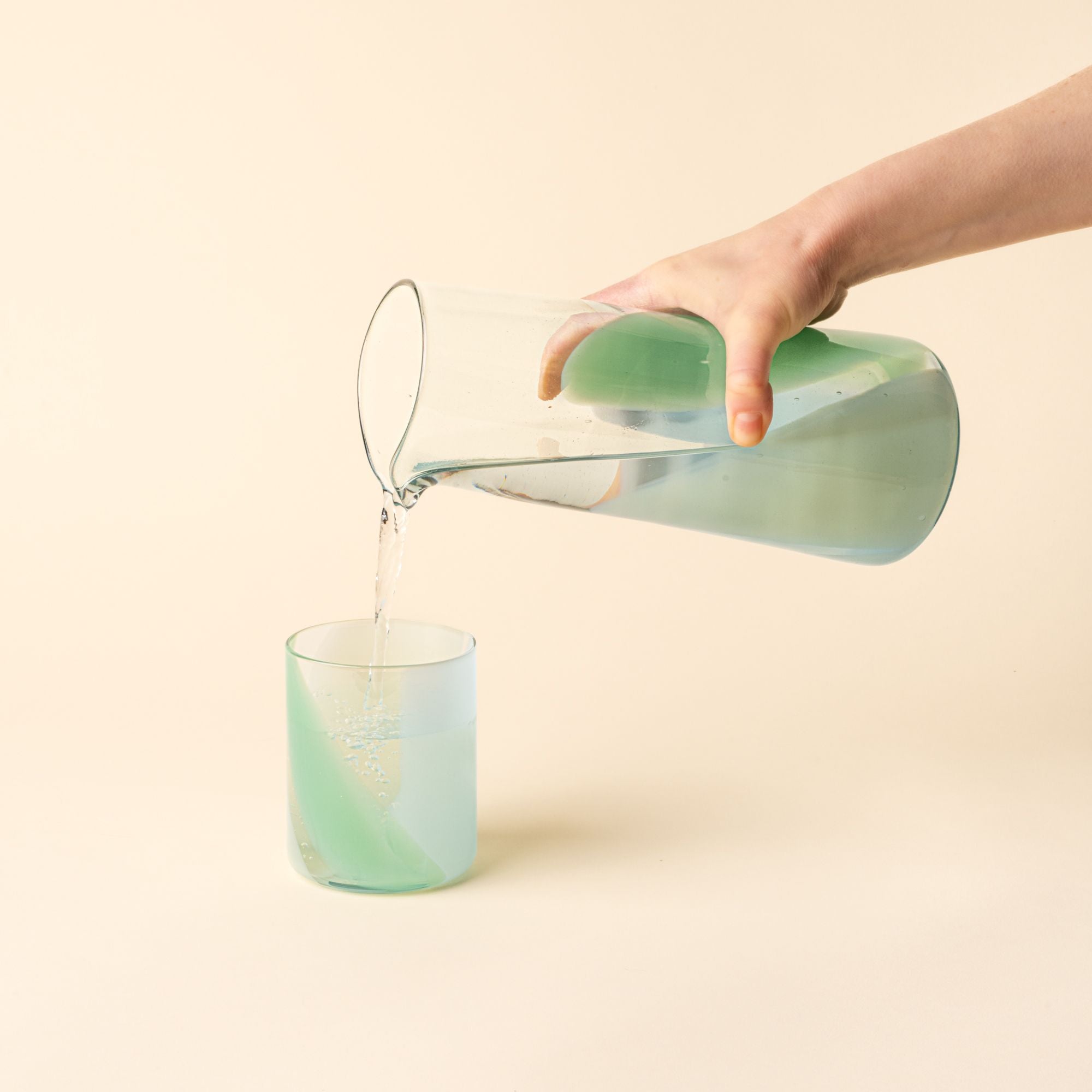 Hand holds a simple glass pitcher featuring a soft pastel green swirl design and pours water into a matching tumbler