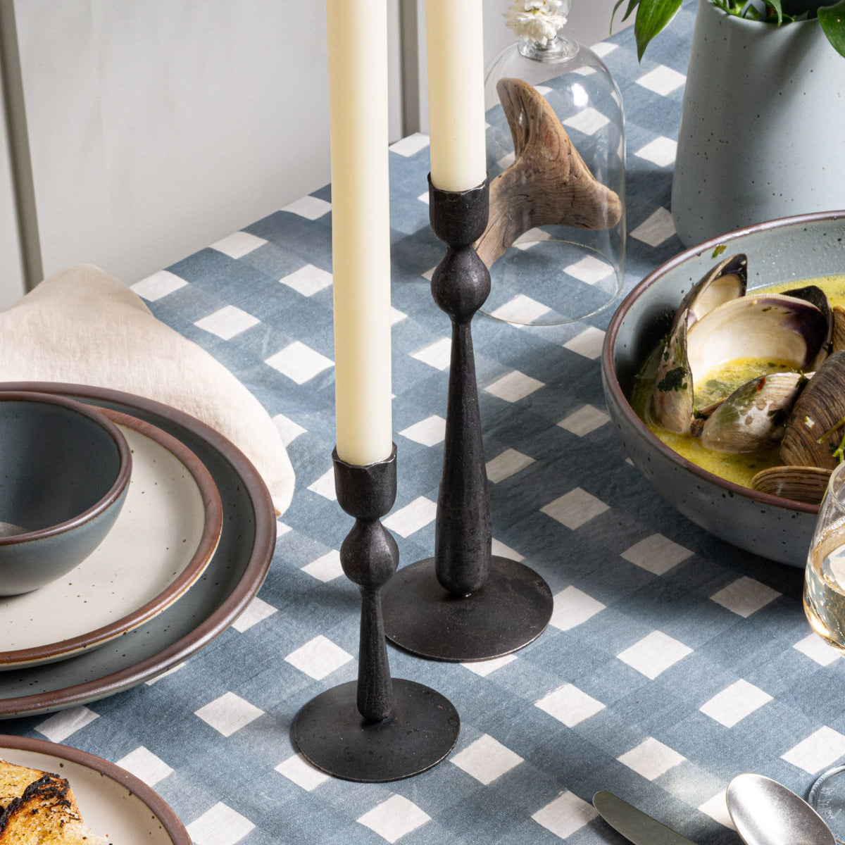 Two forged candle holders with cream tapers on a table with a blue-grey tablecloth with blue-grey and cream ceramic dinnerware