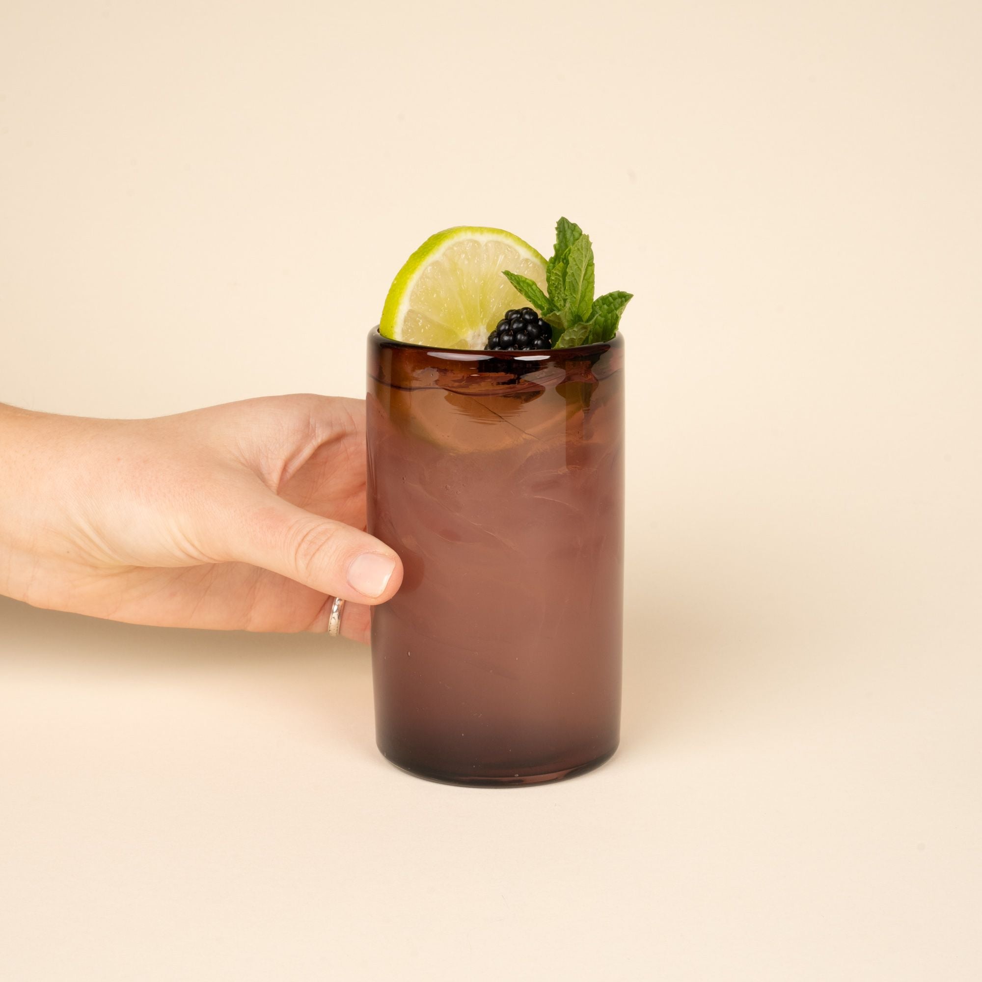 A hand holds a cocktail with a lime, raspberry, and mint garnish in a light plum tall transparent drinking glass