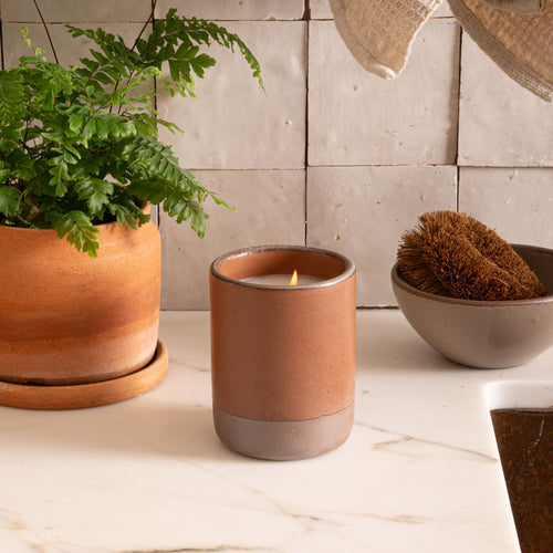 Large ceramic vessel in a cool terracotta color with candle inside on a countertop with a potted plant and dish scrub.