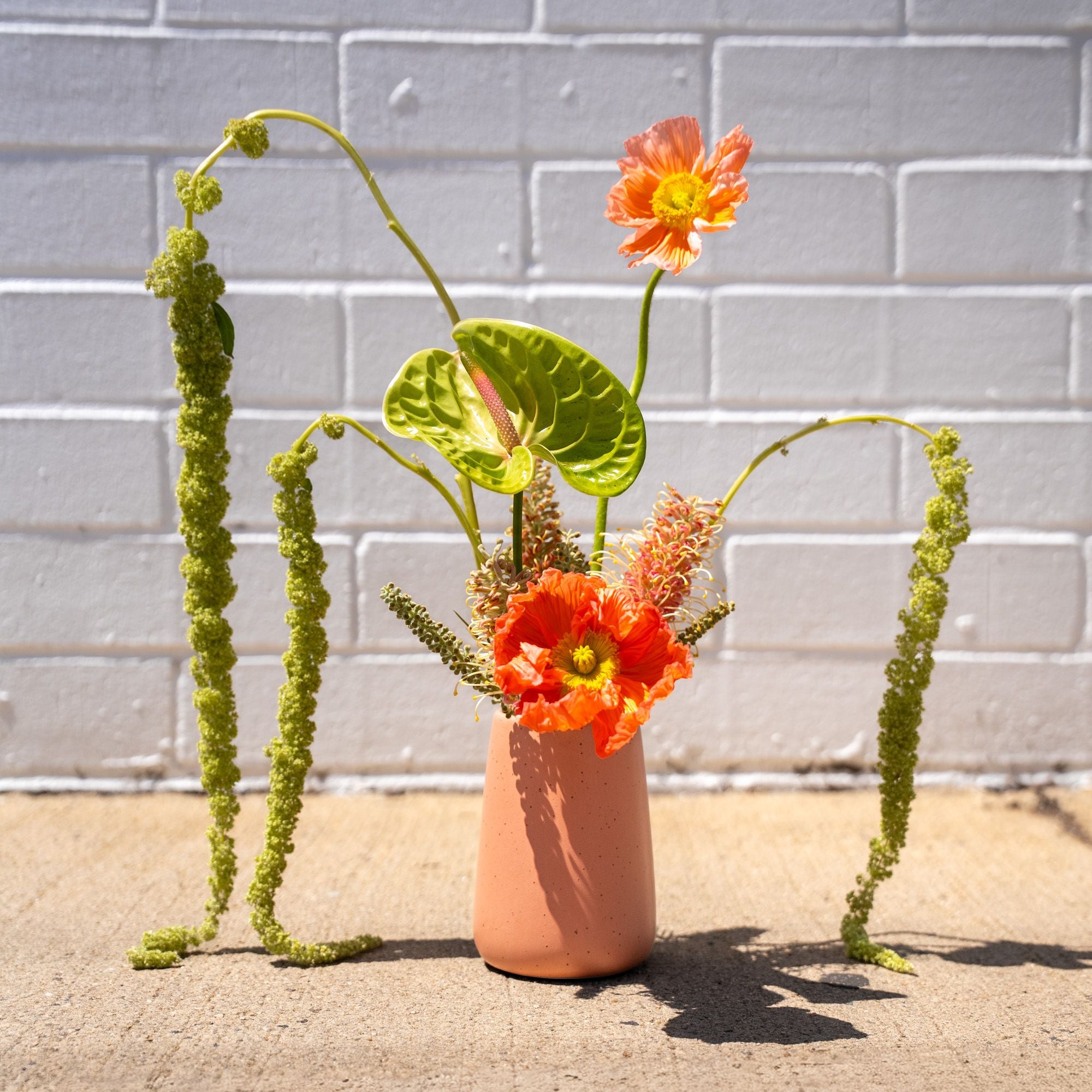 A terracotta-colored ceramic vase with a modern shape, holding a bold arrangement of orange poppies, green anthurium, and trailing amaranthus against a white brick wall.