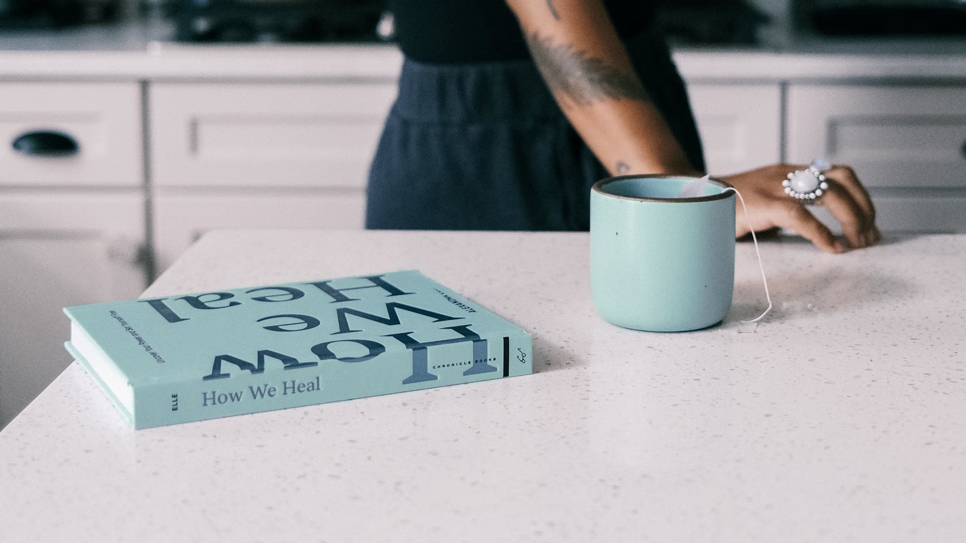 A mint green mug with a tea bag sits on a white speckled countertop next to a book titled "How We Heal". A person with a tattooed arm and large ring leans against the counter in the background.