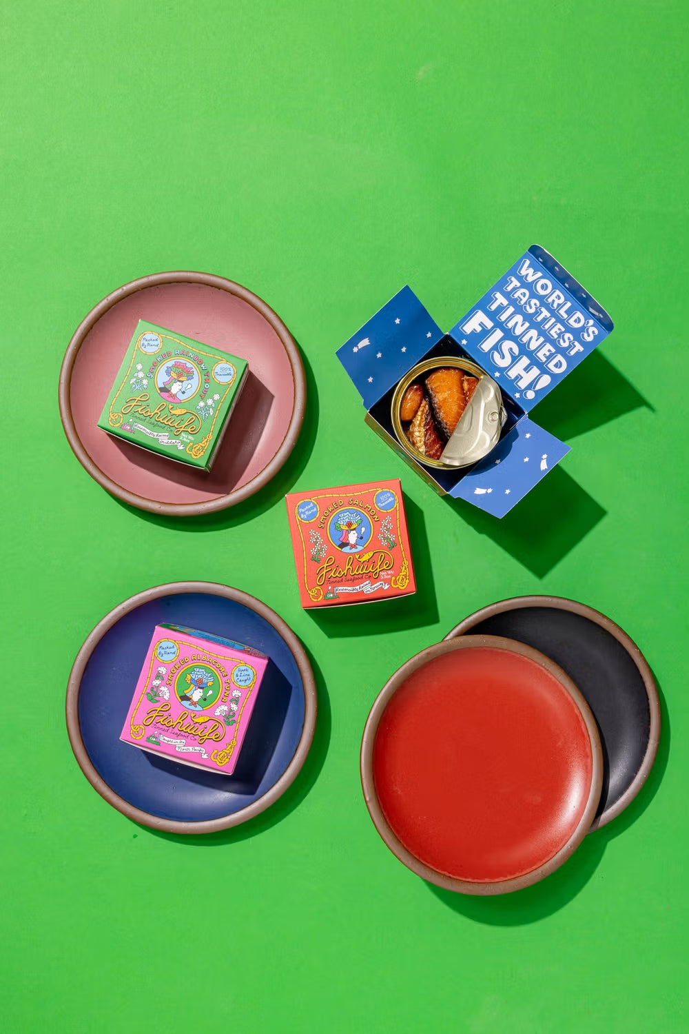 Assorted tins of flavored sardines in colorful packages placed on ceramic plates in various glazes, arranged on a vibrant green background, with one tin opened to reveal the fish inside.