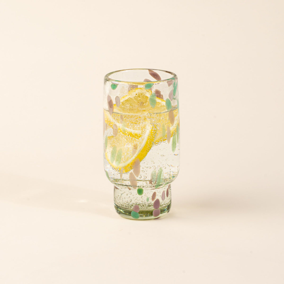 A tall clear glass with pastel green and pink speckled patterns, filled with water and lemon slices.