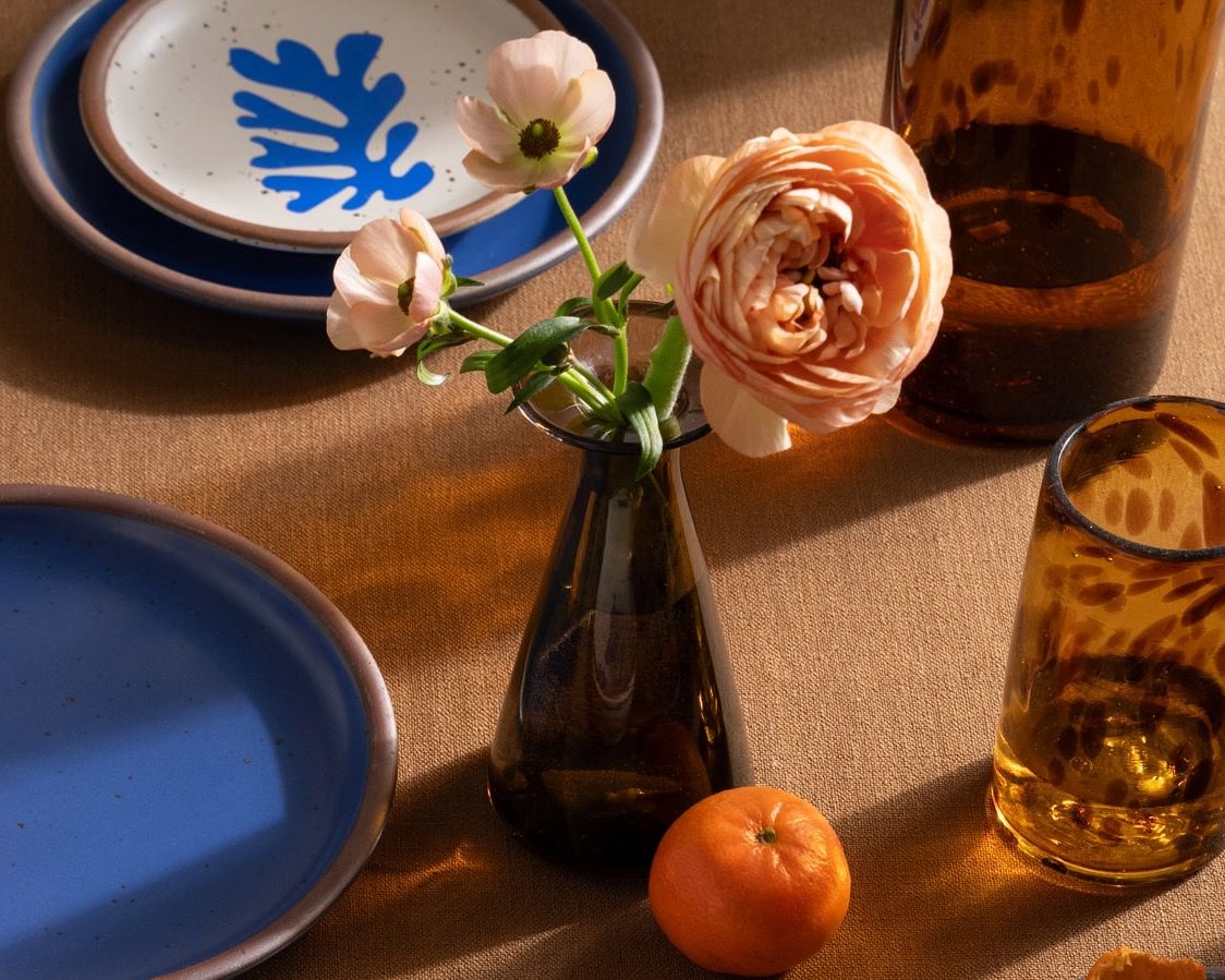 A table setting featuring a blue-rimmed plate with a botanical design, a small amber vase holding pink flowers, an amber glass, and a clementine on a brown tablecloth.