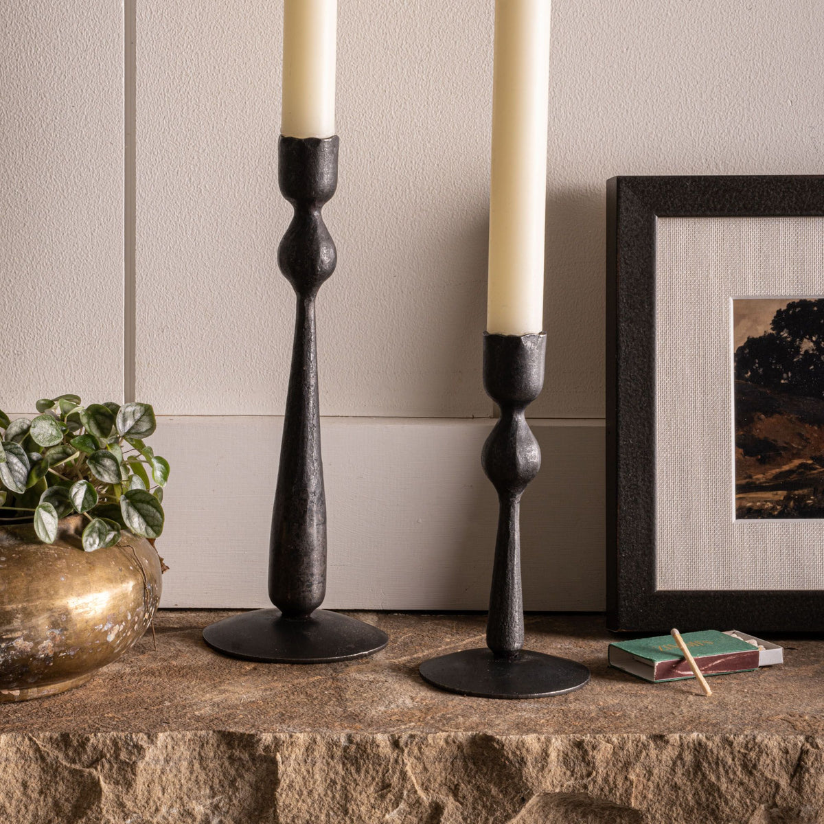 Two forged iron candleholders, one short, one taller, with a cream taper on each, on a mantle.