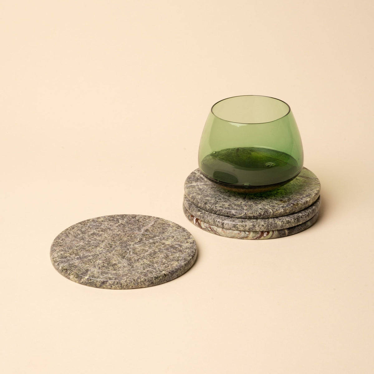 Four round stone coasters with natural green, gray, and brown veining, stacked with one holding a green glass cup.