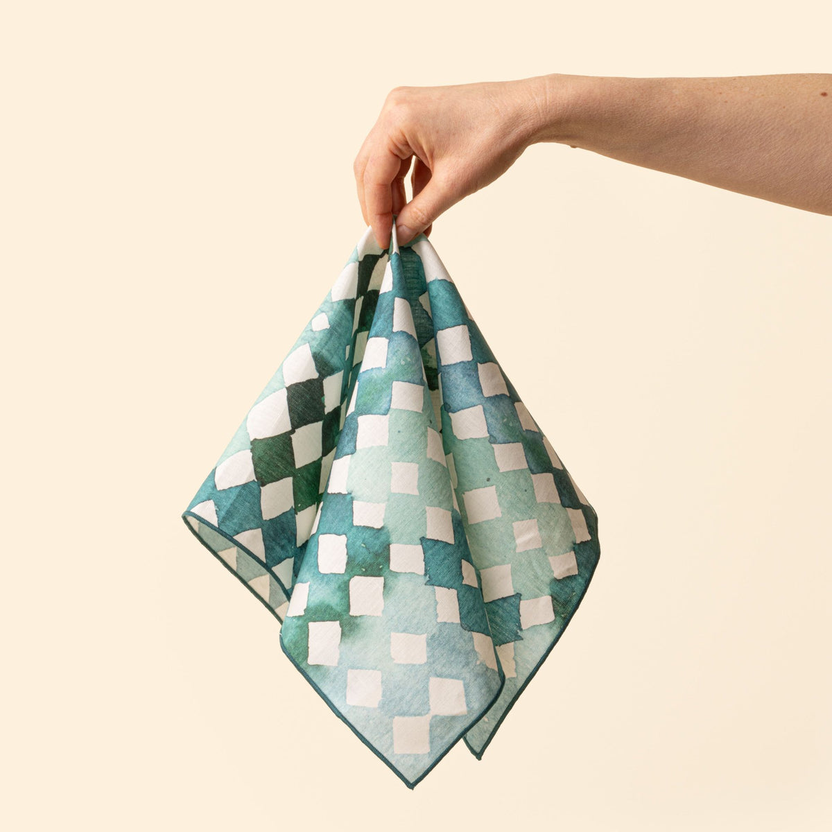 A hand holds out a watercolor diamond-patterned cloth napkin in warm green and teal tones