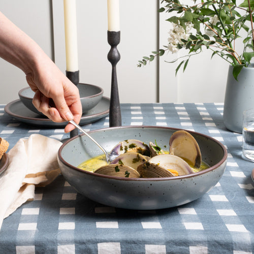 On a table, a hand holds a spoon to scoop from a large shallow bowl in a blue-grey color, filled with a clam dish.