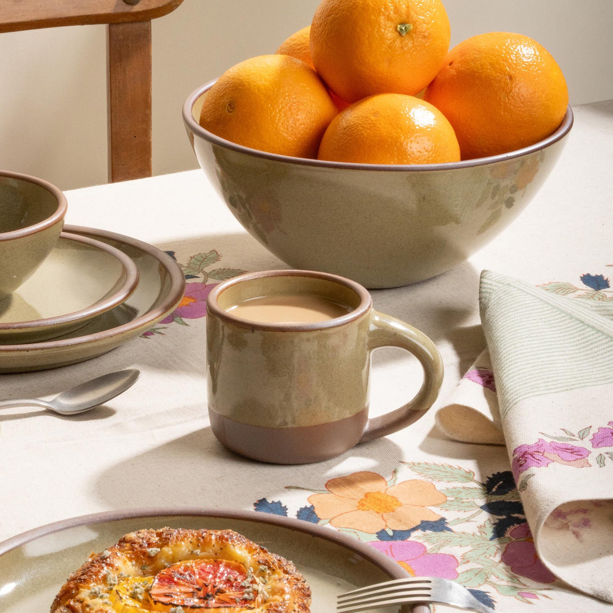 Small stoneware grey-green mug with coffee, bowl of oranges, and breakfast plates on a floral tablecloth.