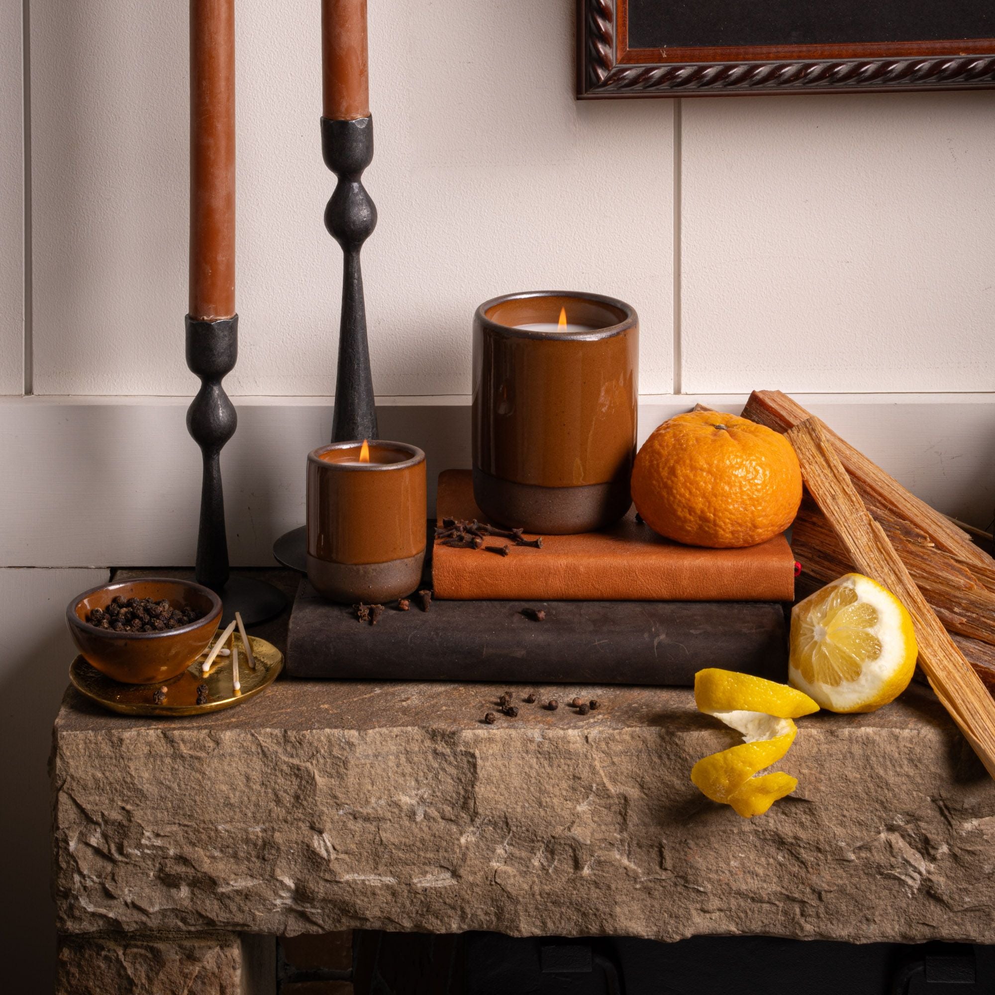 Lit candles in glossy brown ceramic vessels on a stone mantel with citrus, spices, and wood accents for a cozy display.