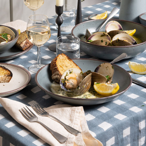 A clam dish and toast sits in a shallow bowl-plate in a blue-grey color, with silverware on a blue-grey crosshatch tablecloth