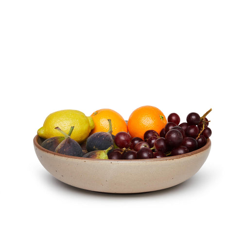 A large shallow serving ceramic bowl in a warm pale brown color featuring iron speckles and an unglazed rim, filled with fruit
