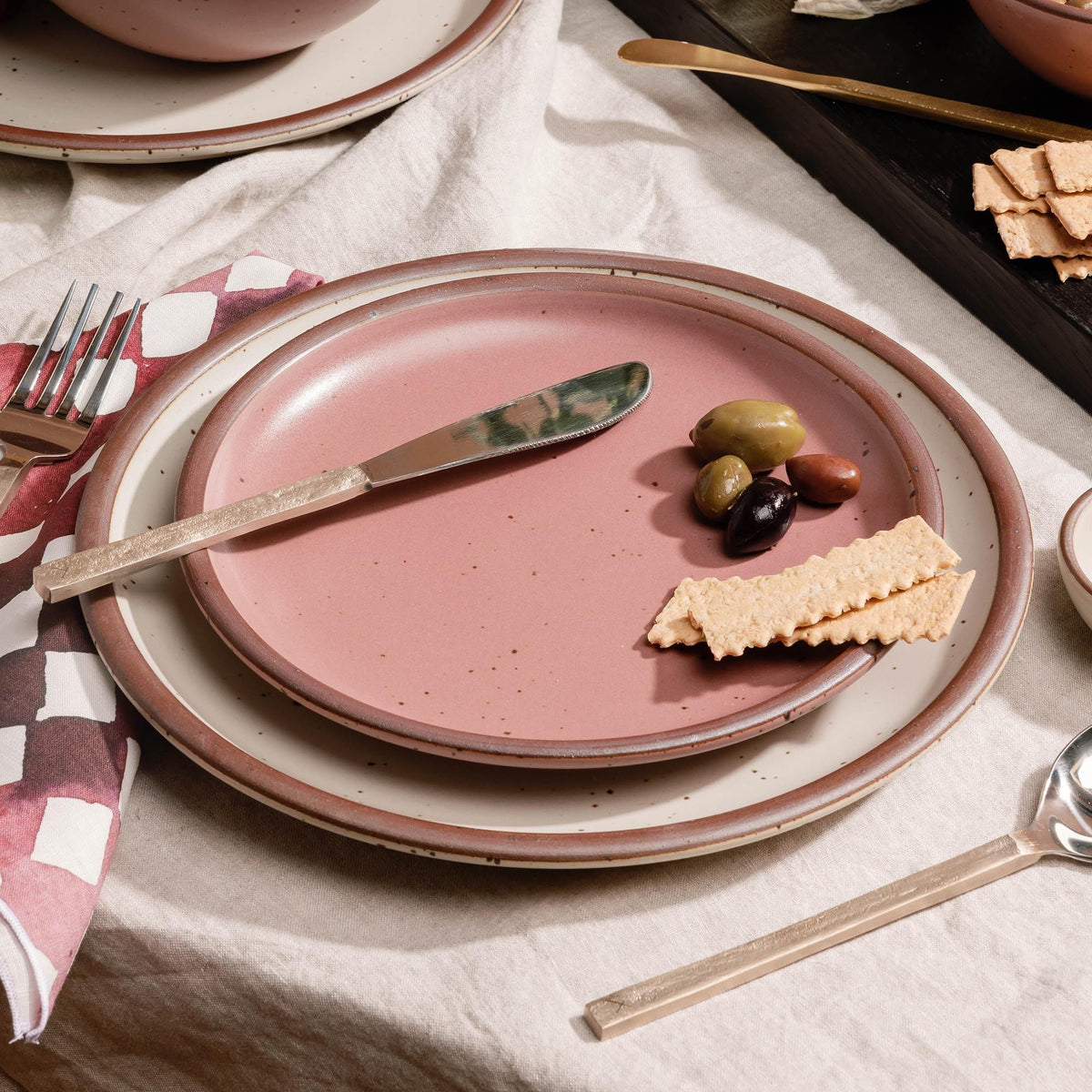 Place setting of dusty mauve pink ceramic side plate and warm off-white dinner plate with a butter knife, crackers, and olives on a white tablecloth with utensils nearby.
