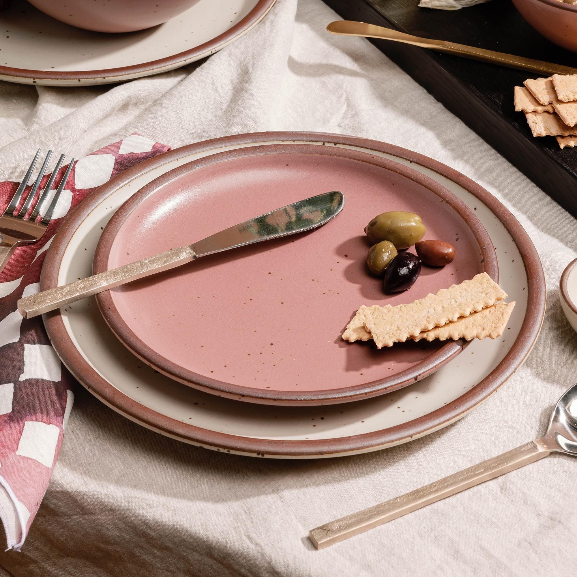 Place setting of dusty mauve pink ceramic side plate and warm off-white dinner plate with a butter knife, crackers, and olives on a white tablecloth with utensils nearby.
