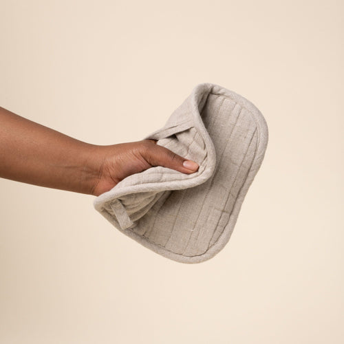 Hand holds out a square quilted linen potholder in a natural color with rounded corners and a small ‘LINEN TALES’ label, against a light background.
