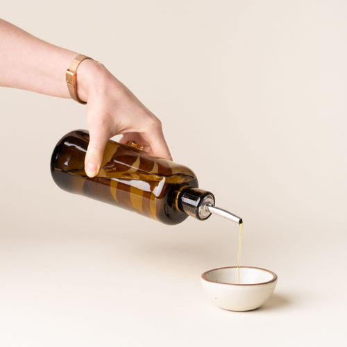 Hand holding an amber glass oil dispenser with a metal spout pouring oil into a small ceramic bowl
