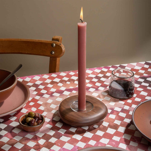 Lit pink taper candle in a wooden holder on a red and pink watercolor diamond tablecloth with tableware and olives.