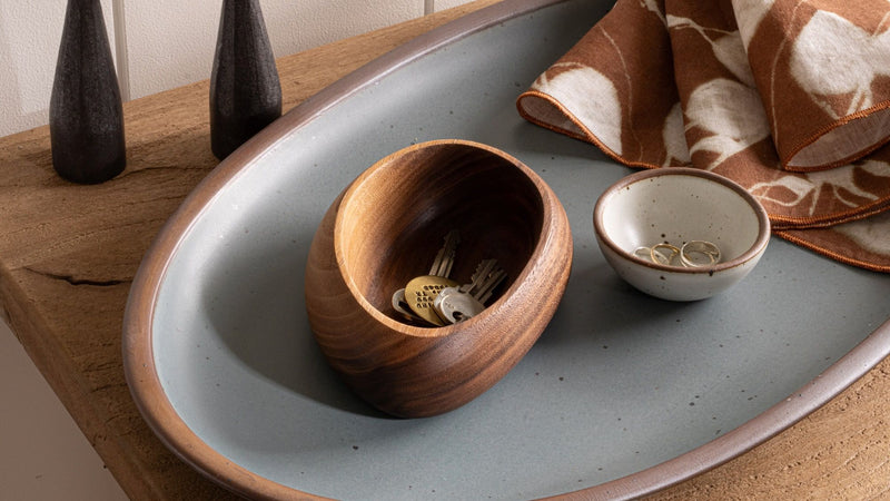 Small round walnut bowl holding house keys, placed on a blue-grey oval platter beside a tiny bowl and printed napkin.