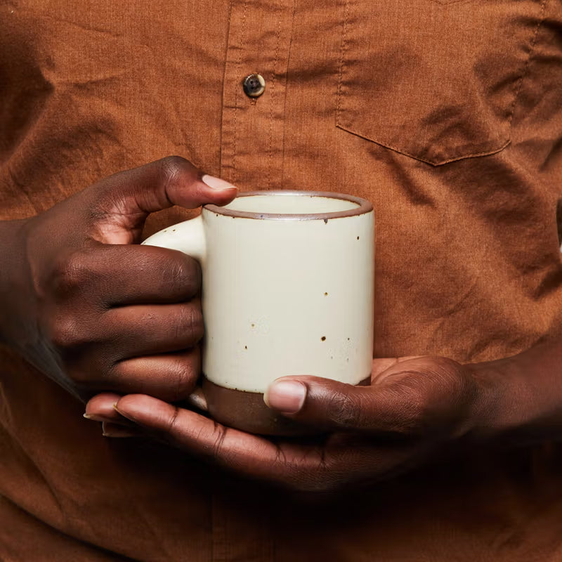 A person wearing a brown shirt holds a cream-colored ceramic mug with a dark rim and base.