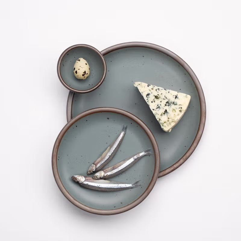 An overhead view a ceramic dinner plate, side plate and tiny bowl in blue-grey colors with styled food on each including fish, cheese, and a quail egg.