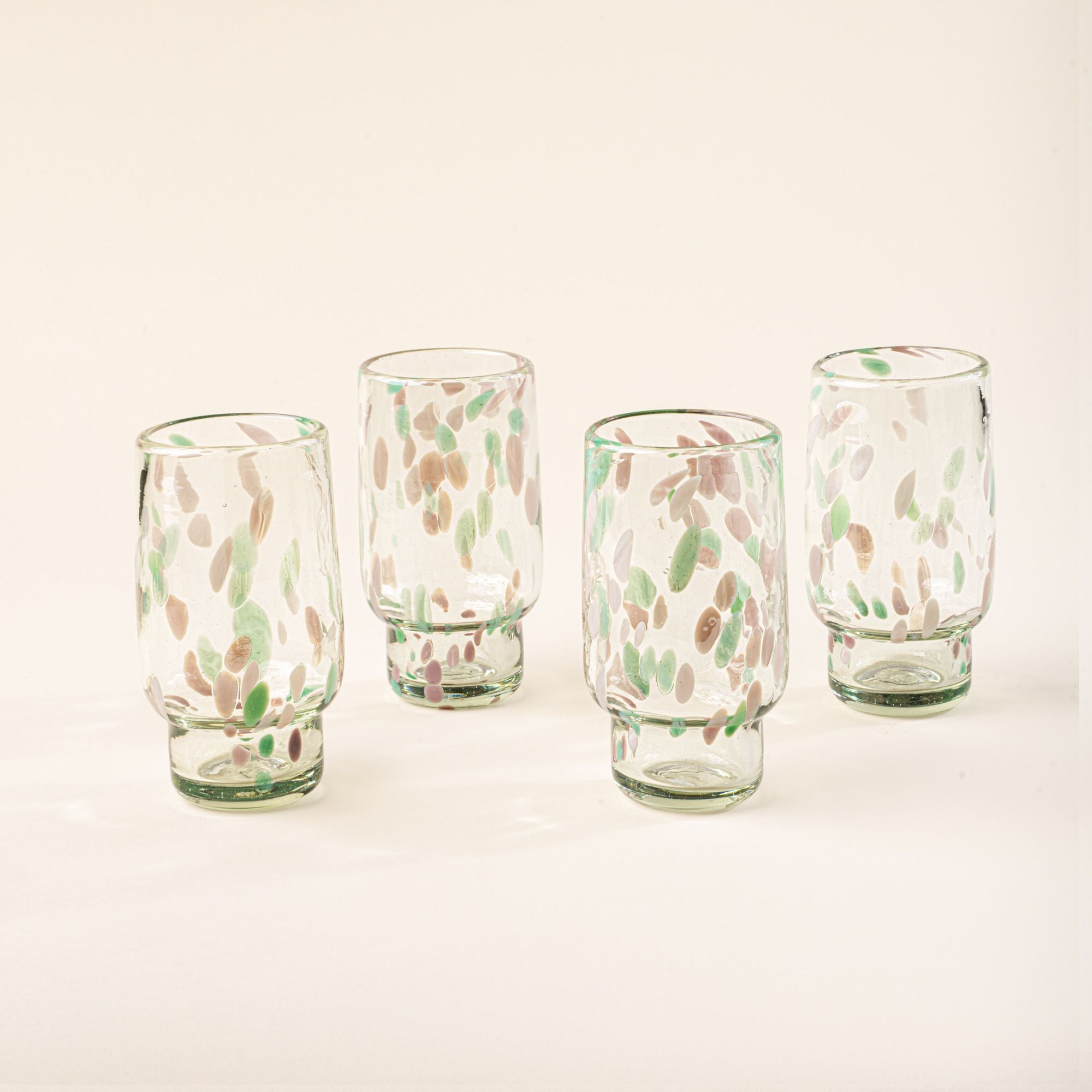 Four tall clear glasses with pastel green and pink speckled patterns arranged in a row on a light background.