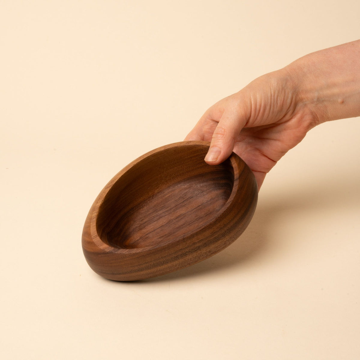 Hand holds out a carved wooden bowl with a smooth, rounded shape and rich brown grain, set against a neutral background.