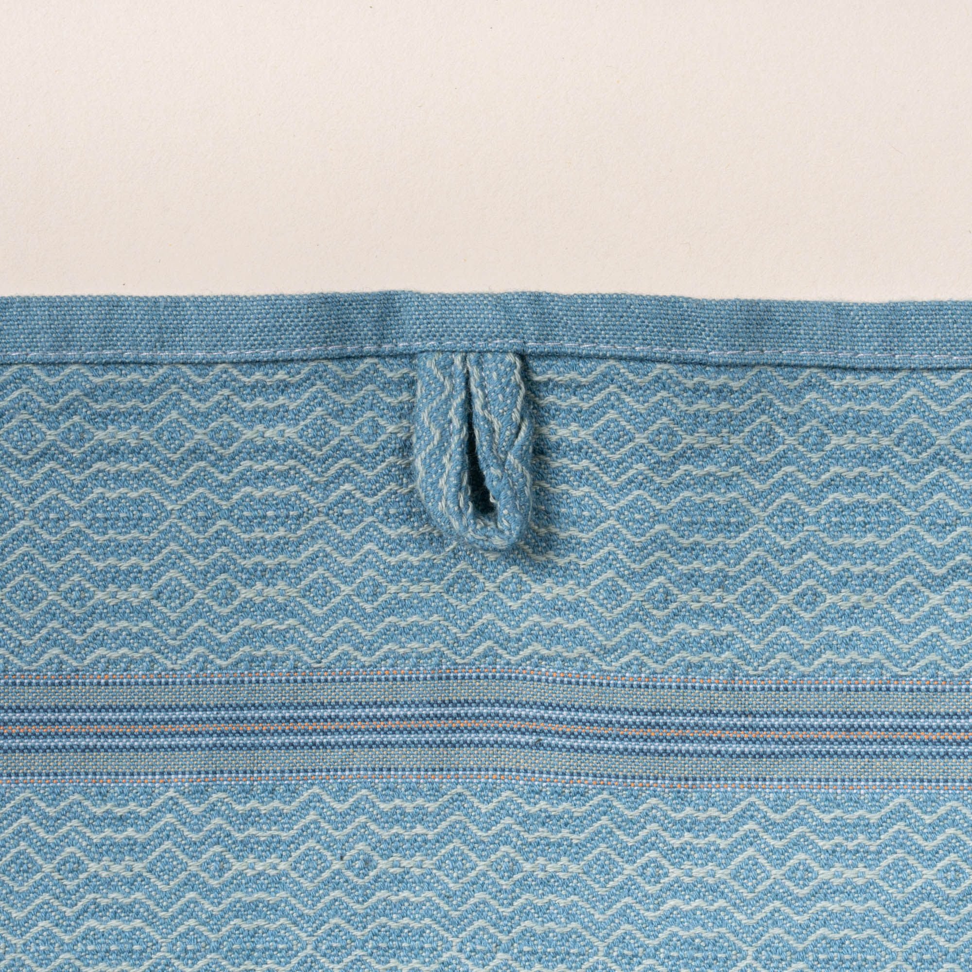 Close-up of a blue woven towel with a zigzag-and-diamond texture, stitched hanging loop, and thin striped band detail.