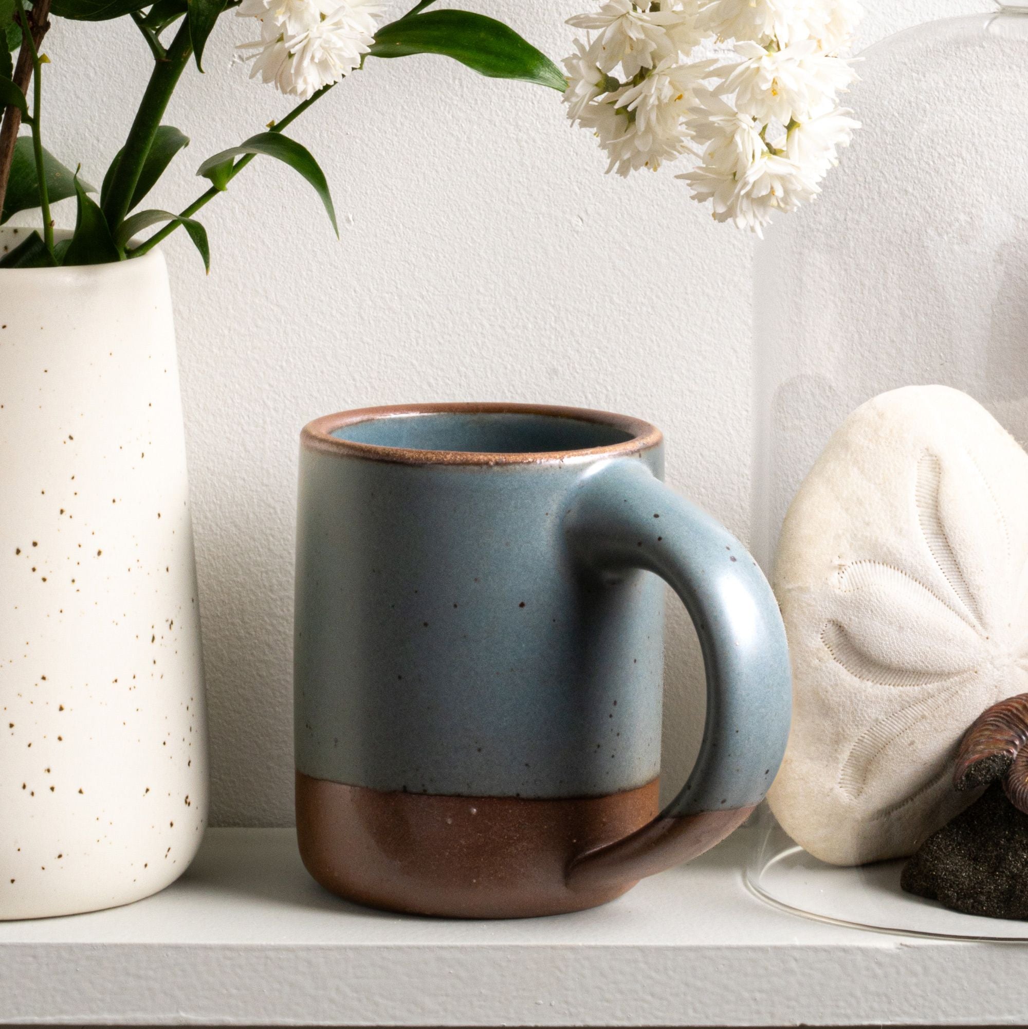 The Mug in a blue-grey color with unglazed base and rim sits on a shelf with a vase of flowers.