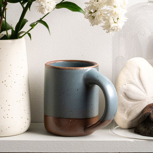 The Mug in a blue-grey color with unglazed base and rim sits on a shelf with a vase of flowers.