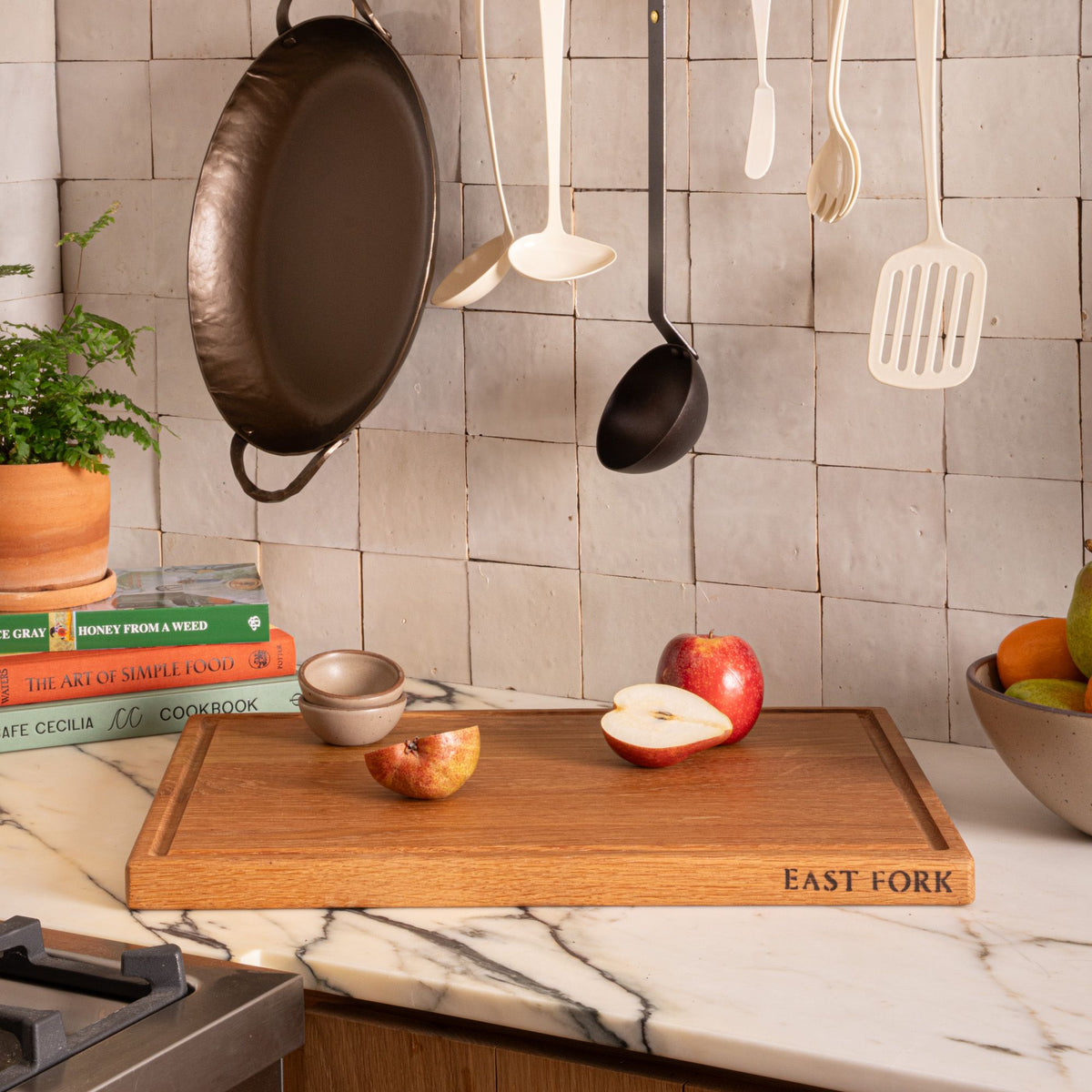 A large wood cutting board with apples and a bowl of fruit on a marble counter, with utensils and a pan hanging on a tiled backsplash.
